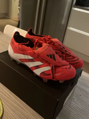 Adidas Predator röda fotbollsskor - Säljer ett par röda Adidas Predator fotbollsskor med vita och svarta detaljer. Skorna har snörning, låg profil och klassisk design med tre vita ränder på sidan. Perfekta för dig som vill dominera på planen med stil. Det är köpta på unisportstore.se kostade 3300kr nypris och är knappast använda pris kan diskuteras