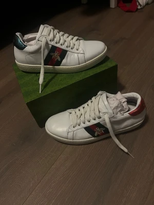 Gucci ace med bi - Snygga vita sneakers från Gucci med klassiska röd- och blå ränder på sidan och en guldbroderad bi. Skorna har snörning, rund tå och detaljer i metallicrött och metallicblått på hälen. Tillverkade i skinn och levereras i originalkartong.