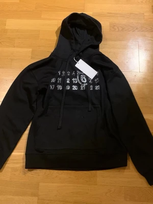 Svart hoodie från Maison Margiela - Säljer en svart hoodie från Maison Margiela med deras ikoniska siffror tryckta i vitt på bröstet. Hoodien har en stor huva med dragsko och en klassisk magficka. Tillverkad i mjuk bomull för skön komfort och stilren look.