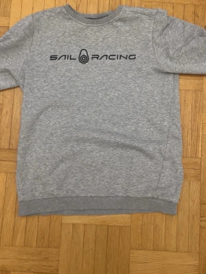 Grå sweatshirt från Sail Racing - Grå sweatshirt från Sail Racing med svart logotyptryck på bröstet och en mindre logga i nacken. Klassisk rund halsringning, ribbade muddar vid ärmslut och nederkant. Perfekt för en avslappnad och sportig stil.
