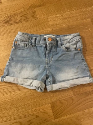 Ljusblå jeansshorts från Lindex - Snygga ljusblå jeansshorts från Lindex med uppvikta benslut och klassisk femficksdesign. Shortsen har normal passform och är perfekta för varma dagar. Tillverkade i mjuk bomull för skön känsla.