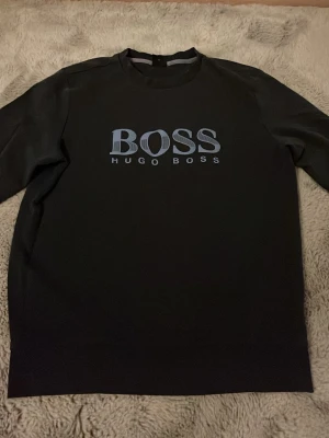 Mörkblå sweatshirt från Hugo Boss - Snygg mörkblå sweatshirt från Hugo Boss med stort BOSS-tryck i ljusblått på bröstet. Tröjan har rund halsringning och ribbade muddar vid ärmslut och nederkant. Perfekt för en stilren och avslappnad look.