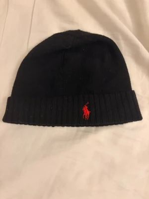 Svart mössa från Polo Ralph Lauren - Snygg svart mössa från Polo Ralph Lauren med uppvikt kant och röd broderad logga framtill. Klassisk och enkel design som passar till det mesta. Tillverkad i mjukt stickat material som håller dig varm under kalla dagar.