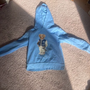 Blå hoodie Polo Bear Ralph Lauren - Säljer en ljusblå hoodie från Ralph Lauren med den ikoniska Polo Bear-trycket på bröstet. Tröjan har huva, långa ärmar och är gjord i mjuk bomull. Perfekt för dig som gillar streetstyle och vill ha något bekvämt men ändå snyggt.