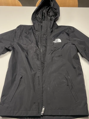 Svart vindjacka från The North Face - Svart vindjacka från The North Face med huva och dragkedja framtill. Jackan har två snedställda fickor med dragkedja och logga på bröstet samt ryggen. Tillverkad i slitstark polyester, perfekt för blåsiga dagar. Använd ca 3 gånger, växt ut den..