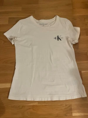 Vit t-shirt från Calvin Klein Jeans - Vit t-shirt från Calvin Klein Jeans med klassisk rund hals och korta ärmar. Diskret CK-logga tryckt på bröstet. Tillverkad i mjuk bomull för en skön och enkel stil som funkar till allt.