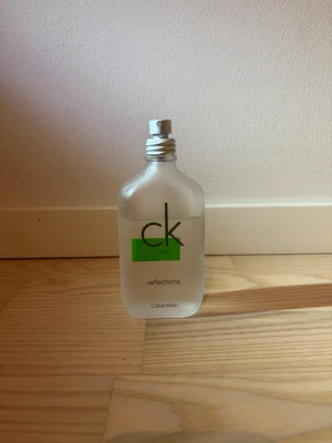 Calvin Klein CK One Reflections EdT - Säljer en stilren parfymflaska från Calvin Klein, CK One Reflections. Flaskan är frostad och genomskinlig med gröna detaljer och har en enkel, modern design. Perfekt för dig som gillar fräscha och unika dofter. Ungefär 3/4 kvar