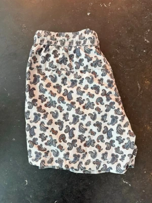 Mönstrade badbyxor med paisley - Säljer ett par snygga badbyxor med paisleymönster i blått och brunt på vit botten. De har elastisk midja med dragsko och sidofickor. Perfekta för stranden eller poolen och ger en chill vibe till sommaren.