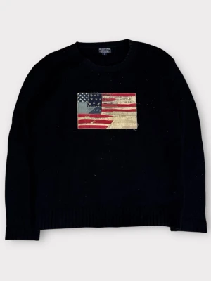 Svart stickad tröja Ralph Lauren - Svart stickad tröja från Ralph Lauren med en stor amerikansk flagga broderad på bröstet. Tröjan har rund halsringning, ribbade muddar och är gjord i ett tjockare garn som ger en klassisk look.