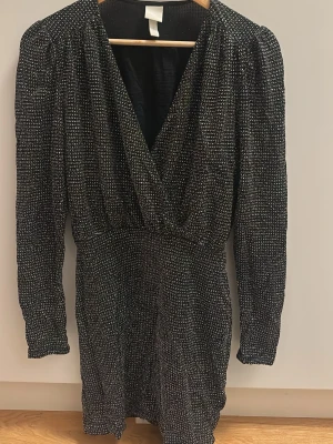 Glittrig svart kortklänning H&M - Säljer en svart glittrig kortklänning från H&M i storlek XS. Klänningen har omlott-v-ringning, långa ärmar och markerad midja. Perfekt för dig som vill sticka ut med lite extra glitter. Materialet är mjukt och stretchigt syntet.