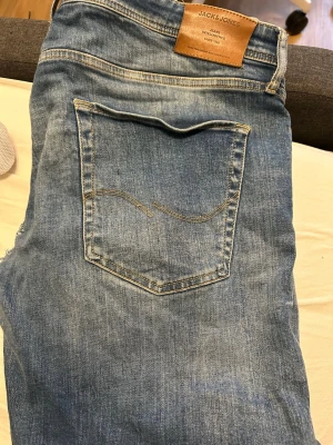 Blå jeans från Jack & Jones - Säljer ett par klassiska blå jeans från Jack & Jones med snyggt slitna detaljer och femficksmodell. Jeansen har normal passform och är tillverkade i bomullsmaterial med en ljus tvätt. Perfekta för en avslappnad och trendig look. Ett hål finns därför säljs de billigt.