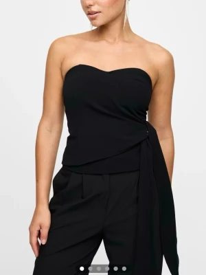 Svart tubtopp med knyt - Stilren svart axelbandslös tubtopp med snygg knyt-detalj i sidan. Toppen har en bandeauformad överdel och är lätt croppad. 