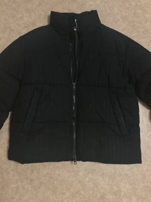 Svart quiltad pufferjacka - Snygg svart pufferjacka med quiltad design och hög krage. Jackan har dragkedja framtill och två sidofickor. Perfekt för kalla dagar när du vill hålla dig varm och samtidigt se stilren ut.