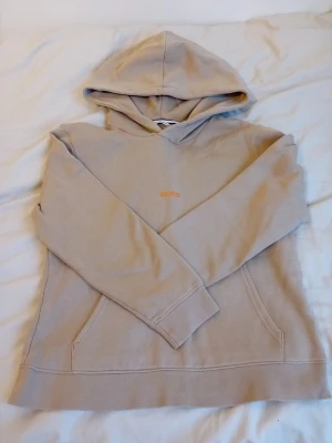 Beige hoodie från Björn Borg - Snygg beige hoodie från Björn Borg med diskret orange logga på bröstet. Hoodien har en klassisk huva och en stor magficka framtill. Tillverkad i mjukt sweatshirtmaterial som är perfekt för chill dagar. Enkel och stilren design som passar till det mesta.