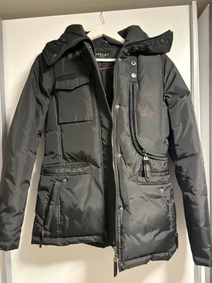 Svart dunjacka från Hollies, stl 38 - Snygg svart dunjacka från Hollies med flera praktiska fickor, hög krage och avtagbar huva. Jackan har dragkedja och knappar framtill samt quiltade detaljer. Perfekt för kalla dagar och har en klassisk, stilren look.