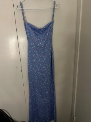 Blommig blå långklänning med smala band - Söt blå långklänning med små blommor i rosa och vitt. Klänningen har smala axelband, lätt draperad urringning och öppen rygg. Perfekt för varma dagar och somriga vibes.