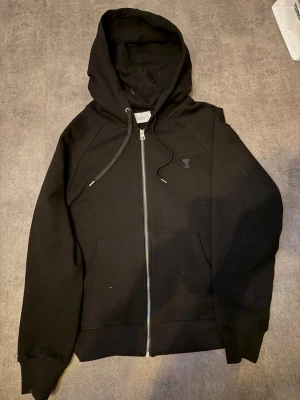 Svart hoodie med dragkedja från Ami - Svart hoodie från Ami med dragkedja framtill, huva med snörning och broderad Ami-logo på bröstet. Tillverkad i mjuk bomull och har två fickor framtill. Perfekt för en chill och stilren look. Den är oversized i luvan