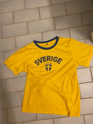 Gul Sverige t-shirt med blå detaljer - Gul t-shirt med blå text 'Sverige' och svensk flagga på bröstet. Blå ribbad krage ger en sportig look. Perfekt för att heja på Sverige! Klassisk passform och kort ärm.