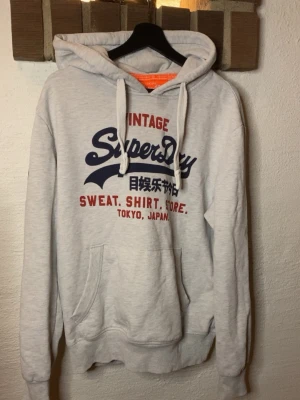 Grå Superdry hoodie med tryck - Snygg grå hoodie från Superdry med blått och rött tryck framtill. Klassisk känguruficka, huva med vita snören och ribbade muddar. Perfekt för en avslappnad streetstyle-look.