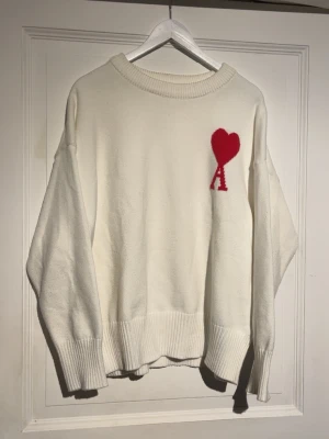 Vit stickad tröja Ami Paris - Vit oversized stickad tröja från Ami Paris med röd hjärtlogga och bokstaven A på bröstet. Tröjan har rund halsringning, breda ribbade muddar vid ärmslut och nederkant samt avslappnad passform. Storlek M/L. Nypris ca 4300kr. Tröjan är äkta, qr-kod plus taggen ligger någonstans i huset (letar). Hör av er vid frågor✌️