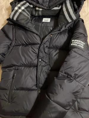 Svart pufferjacka från Burberry - Säljer en svart pufferjacka från Burberry med huva och dragkedja. Jackan har två fickor med dragkedja, ribbade muddar och en klassisk Burberry-rutig insida på huvan. Märket syns tydligt på ärmen. Perfekt för kalla dagar.strolek s och är typ helt ny svarar så snabbt som möjligt