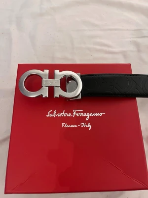 Svart läderbälte Salvatore Ferragamo - Snyggt svart bälte från Salvatore Ferrag sopm har ett präglat mönster i lädret och ett stort, silverfärgat spänne med Ferragamo-loggan. Perfekt accessoar för att lyfta din outfit med en exklusiv touch.