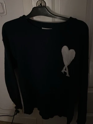 Svart stickad tröja från Ami Paris - Säljer en svart stickad tröja från Ami Paris med deras ikoniska vita hjärta och bokstaven A broderat på bröstet. Tröjan har rund halsringning och långa ärmar. Perfekt för dig som gillar stilrena och trendiga plagg.