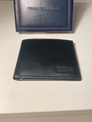 Svart plånbok från Tommy Hilfiger - Stilren svart plånbok från Tommy Hilfiger i slätt skinn. Plånboken har flera kortfack och en diskret logga på framsidan. Perfekt storlek för fickan och levereras i en snygg blå ask med röd söm.