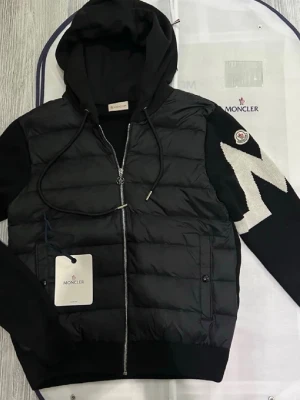 Moncler Cardigan  - Säljer en svart cardigan  från Moncler med huva och dragkedja. Jackan har stickade ärmar med en stor vit M-detalj på ena ärmen och Moncler-logga. Framsidan är quiltad och har fickor med knappstängning. Perfekt för dig som vill ha en snygg och sportig look. Väldigt sällsynt jacka dessvärre priset. Men priset kan såklart diskuteras. Bara att skicka om ni undrar något!