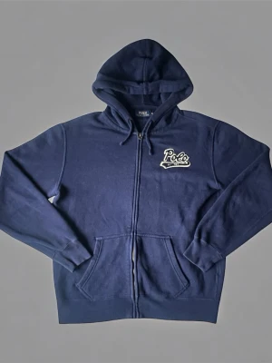 Mörkblå hoodie från Polo Ralph Lauren - Mörkblå hoodie med dragkedja från Polo Ralph Lauren. Broderad Polo-logga med 'EST. 1967' på bröstet. Klassisk huva med dragsko och två fickor framtill. Tillverkad i mjukt bomullsmaterial som är perfekt för chill dagar.
