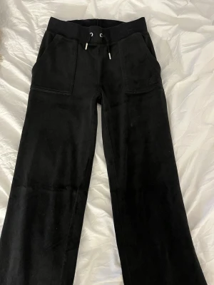 Svarta mjukisbyxor Juicy Couture XS - Juicy Couture mjukisbyxor i svart velour med bred resårmidja och snörning. Byxorna har raka ben, stora fickor fram och bakfickor med knappar. Klassisk JC-logga broderad framtill. Perfekta för chill eller streetwear vibes.