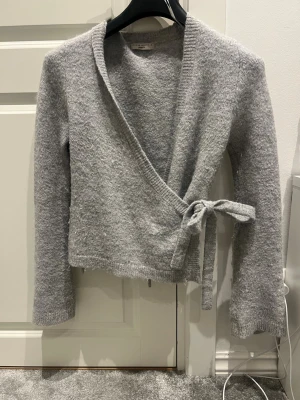 Grå omlottkofta från Gina Tricot  - Mysig grå stickad omlottkofta från Gina  med knytband i sidan. Koftan har lång ärm, ribbade muddar och en mjuk, fluffig känsla. Perfekt att slänga över en topp för en avslappnad look.