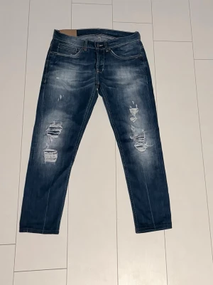 Blå distressed jeans från Dondup - Säljer ett par blå jeans från Dondup med slitna detaljer och snygg tvätt. Jeansen har raka ben och klassisk femficksmodell. Perfekta för dig som gillar en avslappnad och trendig stil.