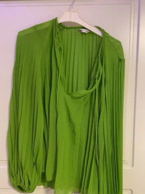 Grön plisserad blus med drapering - Trendig grön plisserad blus med lös passform och draperad halsringning. Blusen har långa, voluminösa ärmar med resår vid ärmslut och är tillverkad i ett lätt, flowy material som ger en snygg siluett.