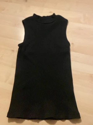 Svart ribbad ärmlös topp - Svart ribbad ärmlös topp från C i storlek 158/164. Toppen har en hög hals och är tillverkad i ett stretchigt material som sitter snyggt mot kroppen. Perfekt att styla med jeans eller kjol för en clean look.