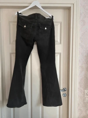 Gråa True religon Jeans! - Snygga gråa True religion jeans med dekorativa ficklock och knappar bak. Jeansen har en klassisk femficksmodell och är tillverkade i ett mjukt jeansmaterial. Perfekta för dig som gillar en retroinspirerad look med utsvängda ben. Helt nytt skick endast testade 