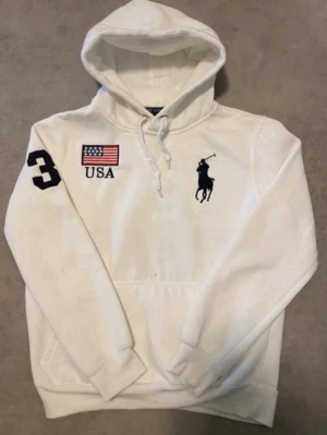 Vit hoodie från Polo Ralph Lauren - Är liten i storlek passar 11-13 åring. Vit hoodie från Polo Ralph Lauren med stor svart polospelare på bröstet, amerikansk flagga och texten USA framtill samt siffran 3 på ärmen. Baksidan har stor siffra 3, UNITED STATES och flagga. Klassisk känguruficka och dragsko i huvan.