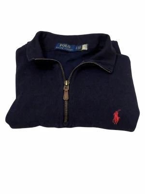 Mörkblå halvzip från Polo Ralph Lauren - Mörkblå tröja med halv dragkedja från Polo Ralph Lauren. Klassisk design med röd broderad logga på bröstet och ribbade muddar. Perfekt för lager på lager och en stilren look.