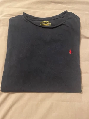 Svart t-shirt från Polo Ralph Lauren - Klassisk marinblå från Polo Ralph Lauren med rund halsringning och den ikoniska röda logotypen broderad på bröstet. Tillverkad i mjuk bomull som känns skön mot huden. Perfekt för en stilren och avslappnad look.