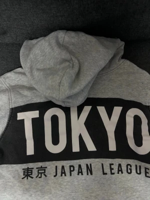 Grå hoodie med TOKYO-tryck / svart hoodie - Snygg grå hoodie med stor svart panel och vitt TOKYO-tryck på ryggen samt texten 'JAPAN LEAGUE' och japanska tecken. En till svart hoodie båda för 150