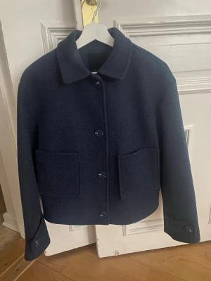 Mörkblå ulljacka från 157, stl S - Säljer en mörkblå ulljacka från 157 i storlek S. Jackan har klassisk krage, stora framfickor och stängs med knappar framtill. Perfekt till höst och vinter för en clean och stilren look.