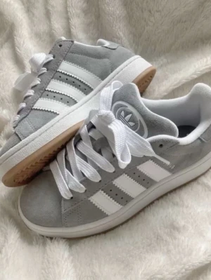 Grå Adidas Campus sneakers - Säljer ett par stilrena Adidas Campus sneakers i grå mocka med vita detaljer och klassiska tre ränder på sidan. Skorna har vit sula och vita skosnören, samt gummisula undertill. Perfekta för dig som gillar en clean och sportig look.