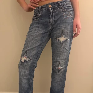 Blå bootcut jeans med stjärndetaljer - Säljer ett par blå jeans med bootcut passform och snygga slitningar i form av stjärnor på benen. Jeansen har dekorativa broderier på fickorna och coola kontrastsömmar. Perfekta för dig som vill sticka ut lite extra! Materialet är klassisk jeans i bomull. Jeansen är vintage och lågmidjade! Skulle säga att dom är storlek S/M 