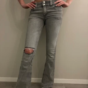 Grå bootcut jeans med hål på knät - Säljer ett par grå jeans med bootcut passform och snyggt slitet hål på ena knät. Jeansen har dubbla knappar i midjan, fickor med lock och knapp bak samt klassisk femficksdesign. Materialet är jeans och färgen är ljusgrå. Jeansen är i storlek 170 men passar modellen som är 18 år och 175cm