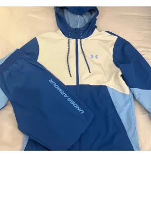Under Armour tracksuite - Snyggt träningsset från Under Armour med jacka och byxor i olika blå nyanser och vitt. Jackan har huva med dragsko, hel dragkedja och logga på bröstet. Byxorna har resår i midjan och logga längs benet. Perfekt för sport eller chill.