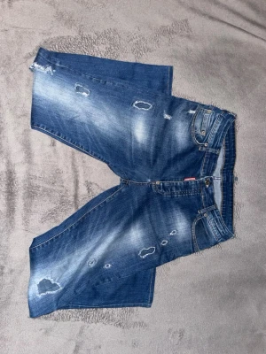 Blå distressed jeans Dsquared2 - Säljer ett par blå jeans från Dsquared2 med slitna detaljer och ljusa tvättade partier. Klassisk femficksmodell med knappgylf och läderpatch bak i midjan. Perfekta för dig som gillar en avslappnad och trendig stil.