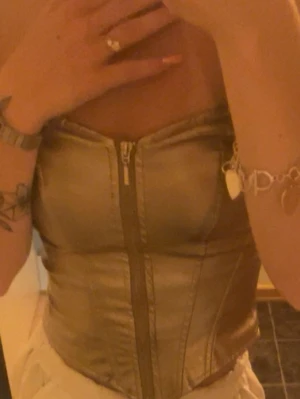Guldig korsett med dragkedja - Snygg guldig korsett med glansig finish och markerade sömmar. Korsetten är croppad och har en dragkedja framtill som ger en cool look. Perfekt att styla till olika outfits för en trendig vibe.
