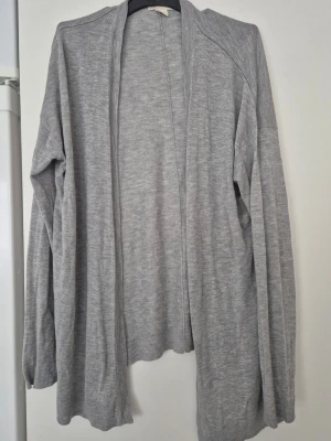 Kofta - Mysig ljusgrå kofta från H&M BASIC i storlek S. Koftan är öppen framtill, har lång ärm och är tillverkad i ett mjukt, tunt stickat material. Perfekt att slänga över en t-shirt eller linne för en chill look.