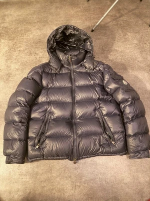 Svart Moncler dunjacka med huva - Säljer en svart dunjacka från Moncler med huva och två snedställda fickor med dragkedja. Jackan har Moncler-logga på ärmen och quiltad design. Perfekt för kalla dagar och har en riktigt puffig look som sticker ut. Storlek 3 och kvitto finns!!