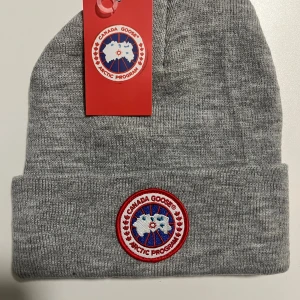Grå mössa från Canada Goose - Säljer en stilren grå mössa från Canada Goose i 100% bomull. Mössan har en klassisk ribbstickad design och är prydd med det ikoniska Canada Goose-märket framtill. Perfekt för kalla dagar och snygg till alla outfits.